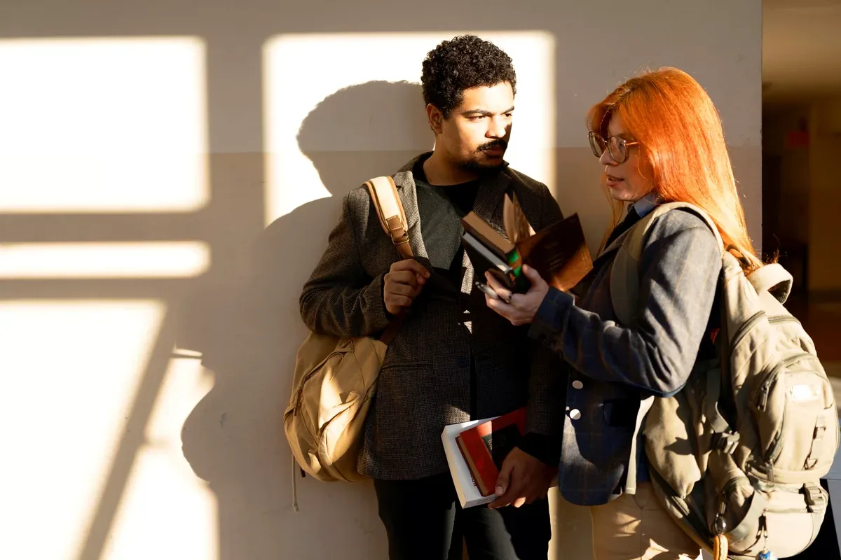 Dos estudiantes con mochilas y libros que conversan junto a una pared