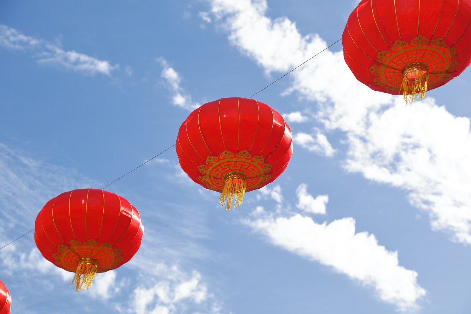 Tres faroles rojos chinos con adornos dorados y flecos colgados de un cable contra un cielo azul con nubes