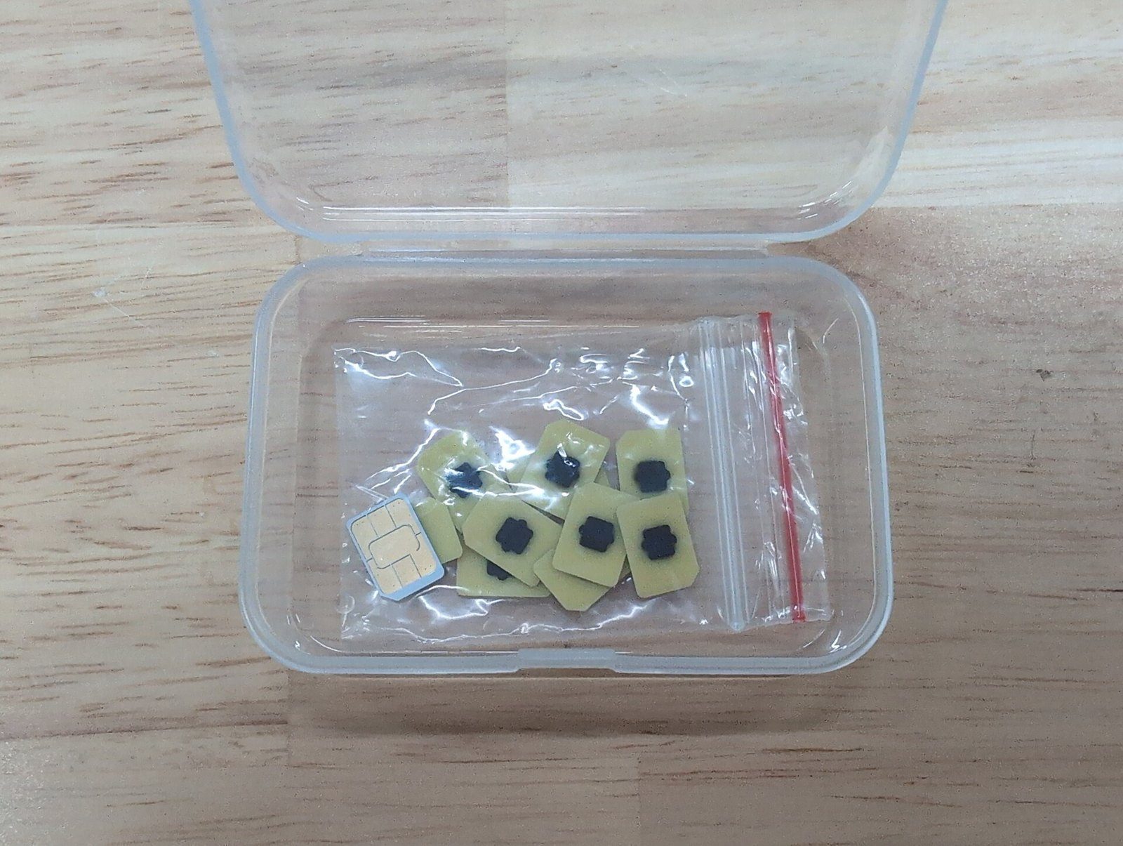 Varios chips eUICC (eSIM) pequeños, de color dorado y negro, junto a una tarjeta SIM, dentro de una cajita de plástico transparente sobre una mesa de madera.